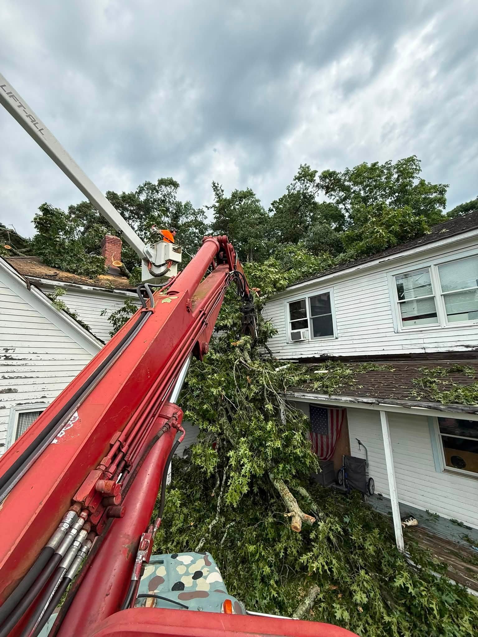 Red crane boom lifting a fallen tree off the damaged roof of a white house